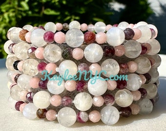 May include: A stack of bracelets made with moonstone, pink, and red beads. The bracelets are layered on top of each other and have a natural, earthy look.