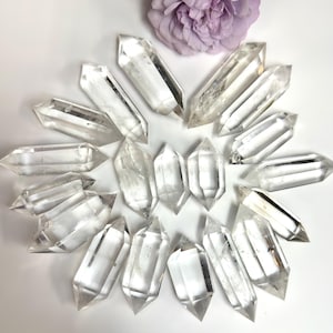 May include: A collection of clear, double-terminated quartz crystal points arranged in a circular pattern around a single light purple flower blossom. Each crystal is transparent, faceted, and features pointed ends on both sides.