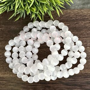 Puede incluir: Una hebra de cuentas de piedra blanca, probablemente selenita, dispuestas en un patr&oacute;n circular sobre una superficie de madera. Las cuentas son lisas y tienen un brillo sutil.