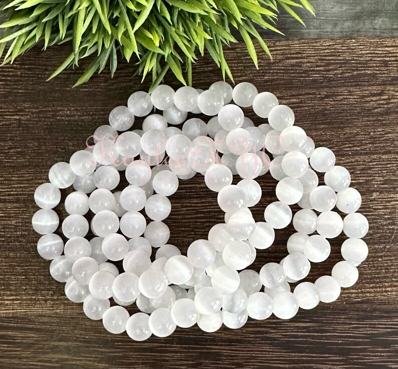 Puede incluir: Una hebra de cuentas de piedra blanca, probablemente selenita, dispuestas en un patr&oacute;n circular sobre una superficie de madera. Las cuentas son lisas y tienen un brillo sutil.