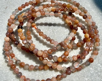 Puede incluir: Múltiples hilos de cuentas de piedras preciosas de piedra solar en tonos naranja, marrón y crema. Las pequeñas cuentas redondas están ensartadas para formar collares. Las piedras preciosas son translúcidas, con variaciones de color y patrón.