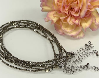 Puede incluir: Un collar de varias hebras con pequeñas cuentas de color gris oscuro y extensiones de cadena plateadas. Una flor rosa y amarilla está en el fondo. El collar es probablemente de pirita.