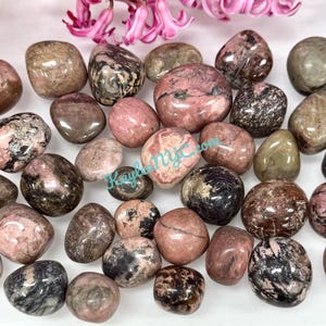 May include: A collection of tumbled pink and black rhodochrosite stones. The stones are smooth and polished, with a variety of shapes and sizes.
