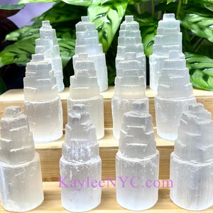 Puede incluir: Una colección de cristales de torre de selenita transparentes, cada uno con un diseño escalonado. Los cristales están dispuestos sobre una superficie de madera, con follaje verde en el fondo. El sitio web KayleeNYC.com es visible.