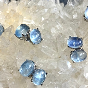 May include: A pair of silver earrings with two oval-shaped moonstone gemstones. The stones are a light blue color and have a shimmery, iridescent finish.