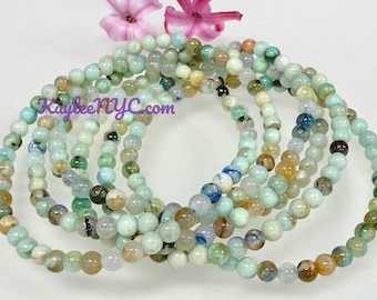 Puede incluir: Un conjunto de seis pulseras hechas con pequeñas cuentas redondas multicolores. Las cuentas son una mezcla de azul claro, verde y marrón. Las pulseras están apiladas unas sobre otras.