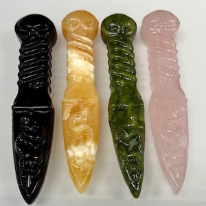 May include: Four carved crystal dagger-shaped objects. The daggers are black, yellow, green, and pink. Each dagger features a skull at the top, a spiral-shaped handle, and a pointed blade. The black dagger has a snake carving.