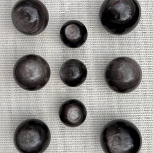 May include: Nine black obsidian spheres of varying sizes are arranged on a light-coloured textured surface. The spheres have a reflective, polished appearance with subtle internal patterns, creating a dark, elegant aesthetic. The spheres range in size from small to large.