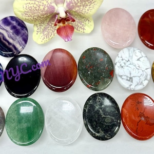 May include: A collection of oval-shaped gemstones in various colors and patterns, including amethyst, rose quartz, and tiger's eye. A light purple orchid flower is in the background. The text "KayleeNYC.com" is visible on some of the stones.