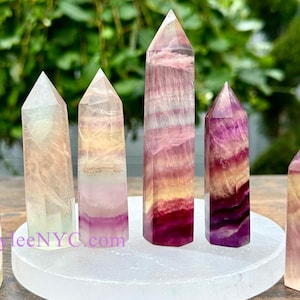 Puede incluir: Seis puntas de cristal de fluorita en varios tonos de rosa, amarillo y blanco, dispuestas sobre una placa de carga de selenita blanca. Los cristales son todos puntiagudos y tienen un aspecto natural y en bruto.