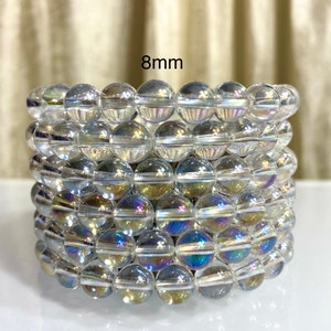 Könnte beinhalten: Sechs übereinander gestapelte, irisierende, klare Quarzkristall-Armbänder, jedes mit 8 mm Perlen. Die Armbänder liegen auf einer weißen Oberfläche. 8mm.
