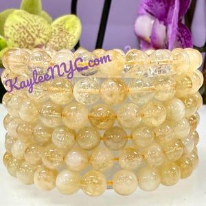 May include: A stack of citrine crystal bead bracelets. The beads are a translucent yellow-orange color and are strung on a yellow elastic cord. The text "KayleeNYC.com" is printed across the top of the bracelets.