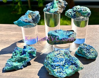 May include: A collection of six rough, natural, blue and green azurite and malachite specimens. The stones are displayed on clear acrylic stands.