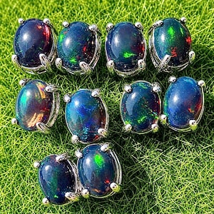 May include: An overhead shot of nine oval-shaped black opal earrings with iridescent flashes of color. Each opal is set in a silver-colored metal frame. The earrings are arranged on a bed of green grass.