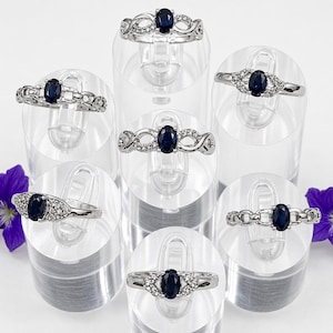 May include: Seven silver rings with oval, dark blue gemstones are displayed on clear ring stands. The rings have various designs, including chain links and infinity symbols, and are accented with small, clear stones. Purple flowers are visible on either side.