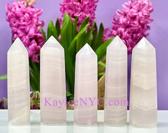 May include: Five pink selenite crystal points arranged in a row on a white surface. The crystals are smooth and translucent, with a slight shimmer. The background is a purple wall with pink flowers.