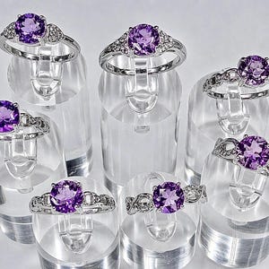May include: Collection of silver rings with large, round, purple gemstones. The rings are displayed on clear, cylindrical stands. Some rings have smaller accent stones. The gemstones are a vibrant shade of purple.