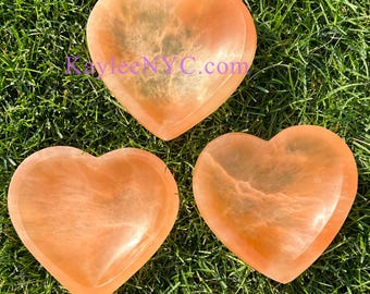 May include: Three heart-shaped bowls made of orange calcite. The bowls are smooth and have a natural, marbled appearance. The bowls are arranged on a bed of green grass.