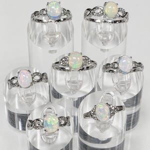May include: A collection of silver rings, each featuring an oval opal gemstone. The rings are displayed on clear acrylic stands. The opals display a range of colors, including white, iridescent, and pastel hues. The bands have decorative detailing.