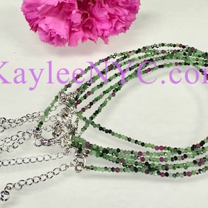May include: Multiple delicate necklaces with green and red beads, each with a silver-colored chain and clasp. The necklaces are displayed on a white surface, with a pink flower in the background. The text "KayleeNYC.com" is visible.