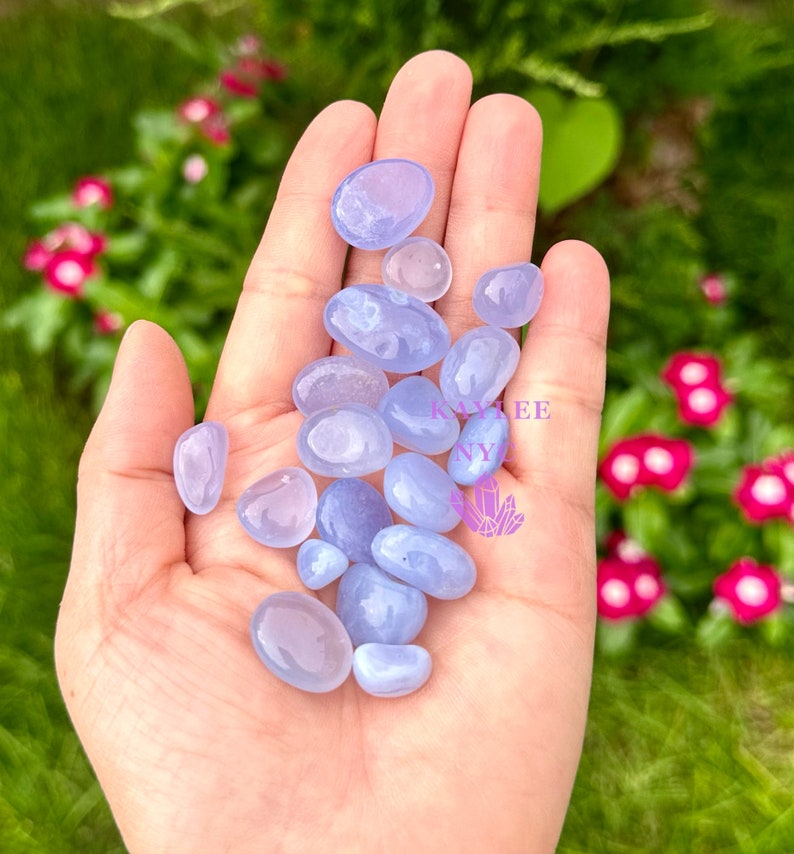 May include: A collection of blue chalcedony tumbled stones in various sizes and shapes. The stones are smooth and polished, with a light blue color and some white inclusions.