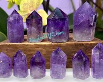 May include: A collection of amethyst crystal points in varying sizes and shapes. The crystals range in color from light lavender to deep purple, with a translucent quality. They are displayed on a wooden block, with the text "KayleeNYC.com" visible.