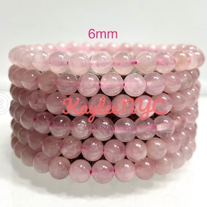 May include: Stack of rose quartz beaded bracelets. Each bracelet is made of 6mm round, light pink beads. The bracelets are stacked on top of each other. The text "6mm" is in the top left corner. The text "Kaylee NYC" is in the middle.