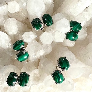 May include: Several pairs of green malachite stud earrings with silver-colored settings are displayed. The earrings have an oval shape and are set against a background of white, crystalline formations.