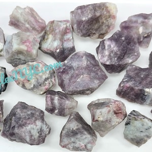 May include: A collection of raw, uncut tourmaline crystals in shades of purple, pink, and gray. The rough stones vary in size and shape, with visible textures and natural formations. The crystals are scattered on a white surface.