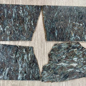 May include: Four irregularly shaped pieces of dark green stone with a textured, fibrous appearance. The stone has streaks of light blue and brown, creating a natural, organic pattern. The pieces are arranged on a light brown wooden surface.