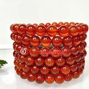 May include: A stack of six carnelian gemstone bracelets, each with round beads. The bracelets are a deep orange-red color and have a glossy finish.