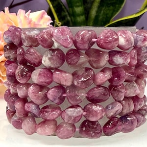 May include: A stack of pink and purple gemstone bracelets. The bracelets are made of polished, irregular-shaped stones strung together. The background includes a soft pink flower and green foliage.