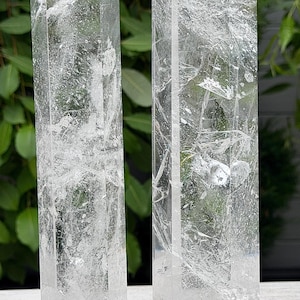 May include: Two clear quartz crystal towers with pointed hexagonal tops. The stones are transparent with visible internal fractures, inclusions, and natural veining throughout the vertical bodies, standing upright with a polished finish.
