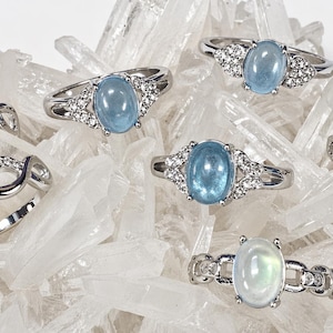 May include: A collection of silver rings with oval-shaped, light blue gemstones. Some rings feature small, clear accent stones. The rings are displayed on a bed of clear crystal formations, creating a sparkling effect.