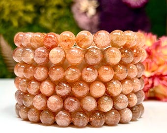 Puede incluir: Una pila de siete pulseras hechas de cuentas de piedra de sol redondas y pulidas. Cada cuenta presenta una base translúcida en tonos melocotón con inclusiones metálicas de color cobre brillante que captan la luz, creando un efecto centelleante.