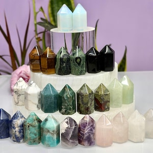 May include: A collection of polished crystal points arranged in rows on a tiered white display. The stones feature various colours and patterns, including blue, green, brown, black, white, purple, and pink, all shaped into smooth, pointed towers.