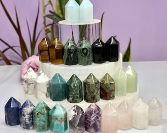 May include: A collection of polished crystal points arranged in rows on a tiered white display. The stones feature various colors and patterns, including blue, green, brown, black, white, purple, and pink, all shaped into smooth, pointed towers.