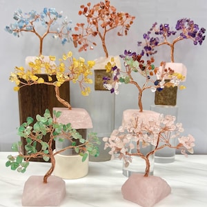 Könnte beinhalten: Eine Sammlung von Edelsteinbäumen mit Zweigen und Stämmen aus Kupferdraht. Die Bäume zeigen verschiedene farbige Edelsteine, darunter blau, orange, lila, gelb, grün und rosa. Jeder Baum ist auf einem rosafarbenen Steinsockel montiert.