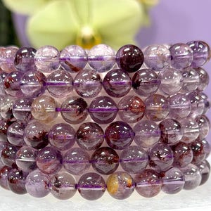Puede incluir: Una pila de pulseras de cuentas de cristal de amatista. Las cuentas son una mezcla de púrpura translúcido y borgoña intenso, con inclusiones visibles. Las pulseras se exhiben sobre una superficie blanca, con un fondo borroso verde y púrpura.
