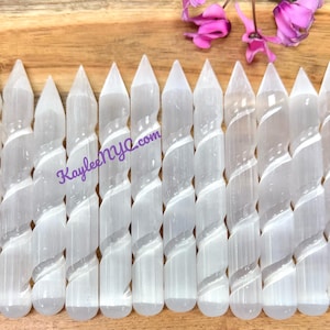 Puede incluir: Una colección de 15 varitas de selenita blanca con un diseño en espiral. Las varitas están dispuestas en una fila sobre una superficie de madera.