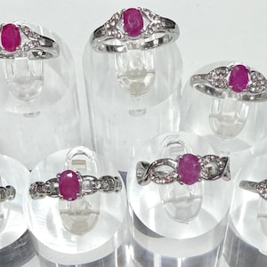 May include: A collection of silver rings, each featuring a vibrant oval ruby as the centerpiece. The rings are adorned with smaller, sparkling accent stones. The rings are displayed on clear, cylindrical stands against a white background.