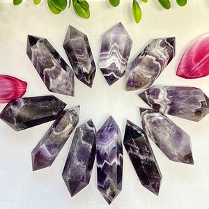 May include: A collection of ten amethyst crystal points, each with a unique purple and white banded pattern. The points are arranged in a circle on a white surface.