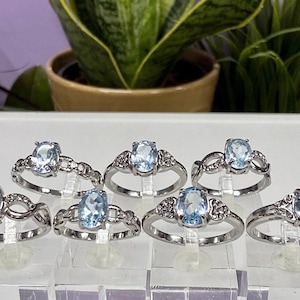 May include: A collection of silver rings with oval, light blue gemstones. The rings are displayed on clear stands, showcasing their intricate designs. The gemstones are the focal point, complemented by the silver bands. The background features green plants.