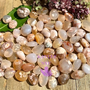 May include: A collection of polished, tumbled stones in shades of beige, cream, and peach. The stones vary in size and shape, with some displaying floral-like patterns. The arrangement is on a wooden surface, with a few green leaves and purple flowers.