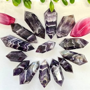 May include: A collection of twelve amethyst chevron double terminated points. The points are a deep purple color with white chevron patterns. The points are arranged in a circle on a white background.