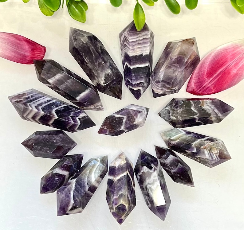 May include: A collection of twelve amethyst chevron double terminated points. The points are a deep purple color with white chevron patterns. The points are arranged in a circle on a white background.