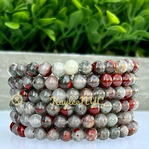 May include: Stack of beaded bracelets. Each bracelet features round beads in shades of gray, red, and white. The bracelets are stacked on top of each other, showcasing the variety of colors and textures. The background is a blurred green.