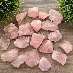 May include: A collection of pink rose quartz crystals, rough and unpolished, with natural variations in shape and size. The crystals are arranged on a wooden surface.