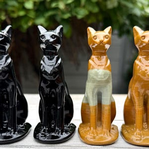 May include: Four ceramic cat figurines standing in a row. The two on the left are solid glossy black, while the two on the right are a warm, glossy amber-brown colour. Each cat is depicted in a seated, upright posture with pointed ears.