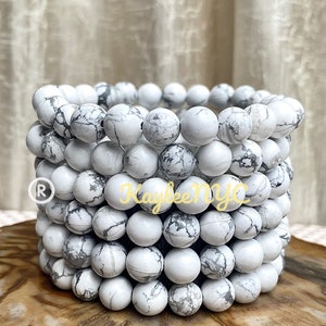 May include: A stack of white howlite bead bracelets with gray veining. The bracelets are made of round beads and are stacked on top of each other.